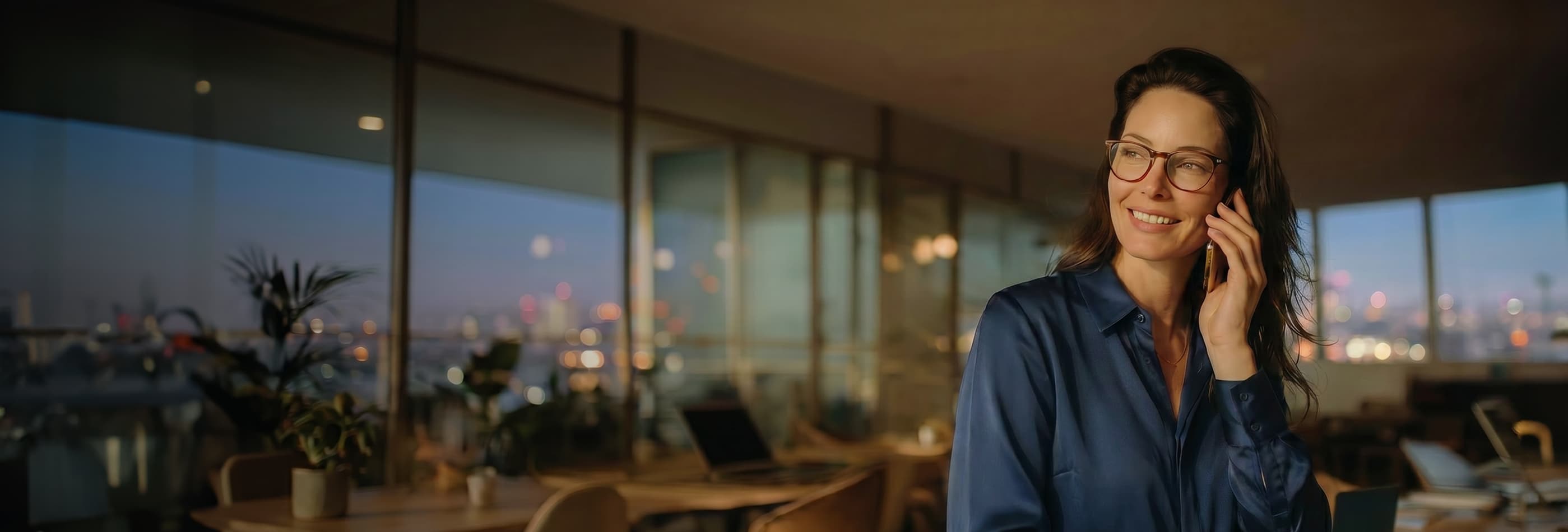 Mulher elegante falando no celular, em um ambiente com luz quente e fundo desfocado.