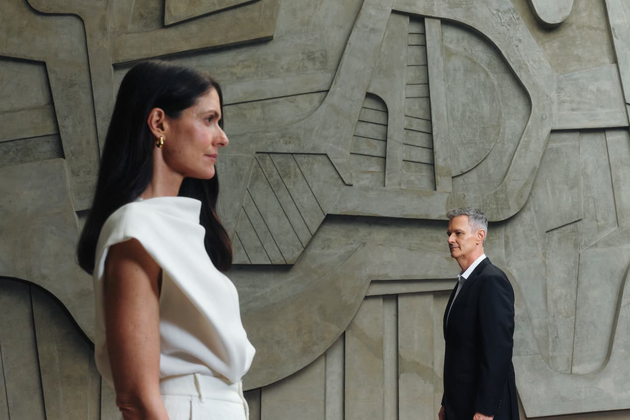Mulher e homem em ambiente corporativo com parede de concreto geométrica ao fundo.