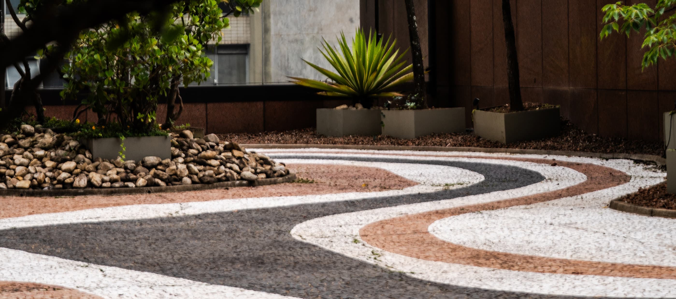 Caminho externo com piso curvo, plantas ornamentais e vasos em área ajardinada.